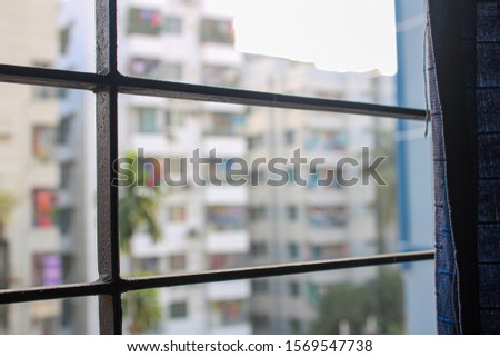 blurred view of high rise buildings from a window while the grill is visible and a part of blue curtain in Elephant Road, Dhaka