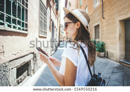 Female hipster tourist with a backpack walking through old town street browsing online city maps application on mobile phone. Travel blogger having vacation in Italy. Tourism and freelance concept.