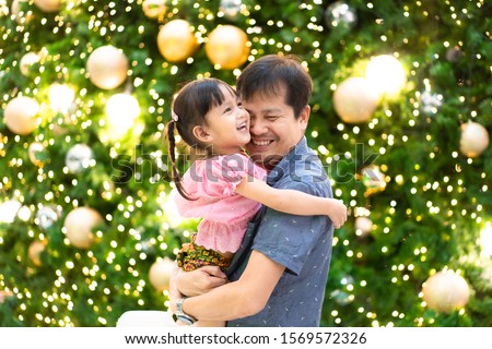 Asian father and cute little daughter are hugging and smiling together with special and happiness moment with the background of green christmas tree and beautiful lighting, authentic family lifestyle.