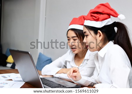 Two cheerful young Asian business women with santa claus hat shopping online together in christmas holiday. Internet of thing concept.