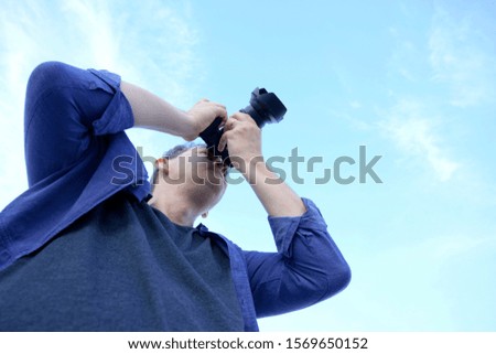 Photographer male in blue shirt holding camera up taking picture over sky background.
