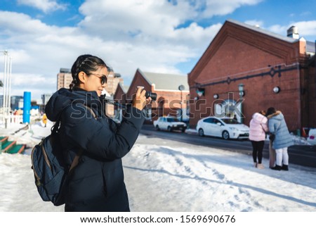 Winter travel concept, Happy traveler asian woman or photographer with backpack and camera against snow relax in red brick warehouse at Hakodate city, Hokkaido, Japan