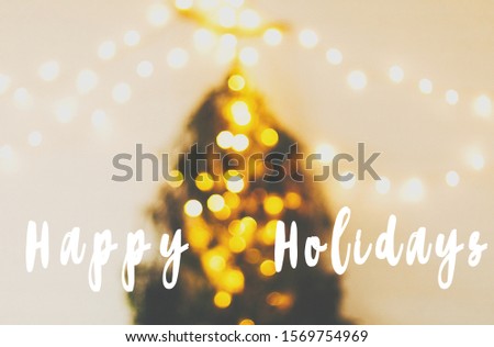 Happy Holidays text sign on abstract christmas tree. Modern christmas tree made of pine branches with golden festive lights hanging on white wooden wall. Seasons greeting card