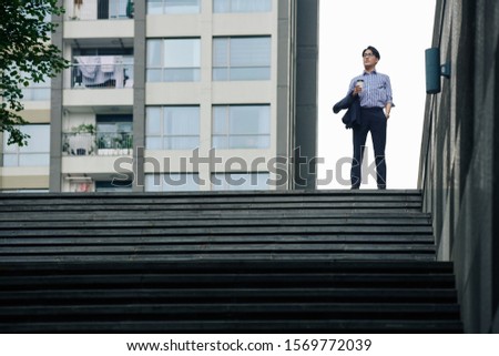 Confident ambitious Asian businessman standing on top of stairs with cup of coffee and jacket in hands and looking forward