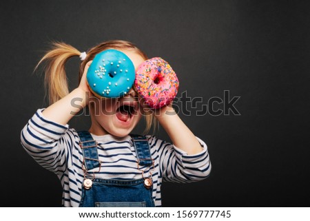 Happy cute girl is having fun played with donuts on black background wall. Bright photo of a children Colored donuts