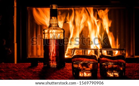 A bottle of whiskey and two glasses with a drink stand on the table against the background of the fireplace where the fire is burning