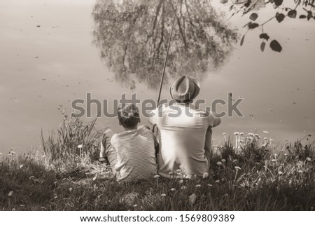 Back view of happy family on summer vacations concept. Father and son fishing together at river bank at scenic landscape background of fresh green grass and blue calm water. Horizontal sepia photo.