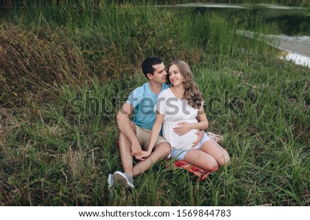 Portrait of a young family expecting a baby. A loving man and a beautiful pregnant blond woman are sitting on a plaid in green grass, hugging and smiling. Pregnancy picture, concept.