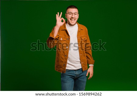 young handsome man feeling successful and satisfied, smiling with mouth wide open, making okay sign with hand against flat background