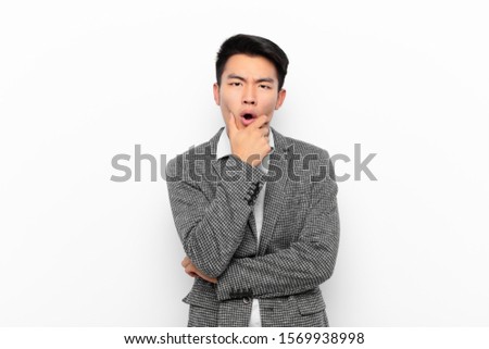 young chinese man with mouth and eyes wide open and hand on chin, feeling unpleasantly shocked, saying what or wow against flat color wall