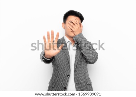 young chinese man covering face with hand and putting other hand up front to stop camera, refusing photos or pictures against flat color wall