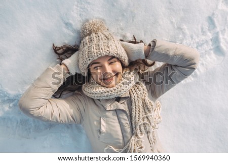 Portrait of a happy smiling young woman laying down on a snow, playing winter games during a sunny winter vacation. Mixed race Asian Caucasian gorgeous girl resting during winter holidays. Female