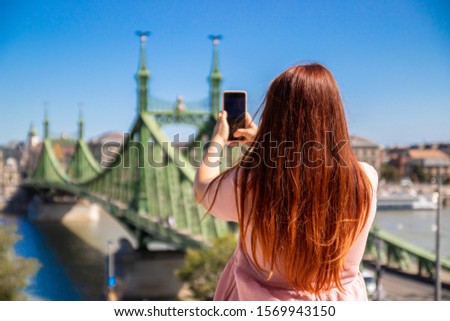 Young red-haired girl in a pink dress makes a photo of the bridge on a smartphone on a sunny summer day