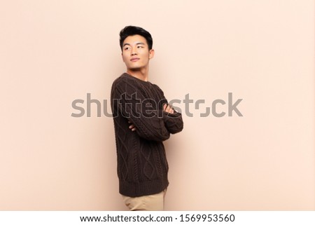 young chinese man smiling gleefully, feeling happy, satisfied and relaxed, with crossed arms and looking to the side against flat color wall