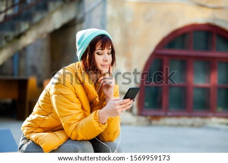 Image of beautiful stylish woman holding mobile phone. Young woman standing at the street and using mobile phone. Woman listening to music with the phone and having fun.