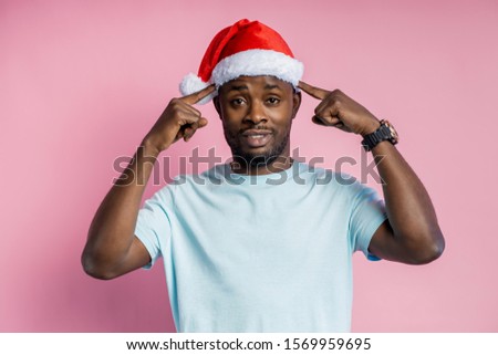 Young calm afro american man with small beard, wearing Santa hat, white t shirt, keeping forefingers on temples, trying to concentrate or remember something, posing over pink background. Body language