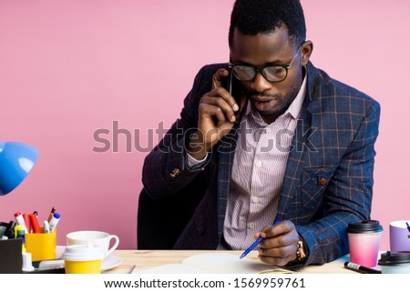 Serious afro american businessman in formal wear, glasses, talking on mobile phone, looking at notepad, holding pen, sitting at desk in office. Job, business concept.