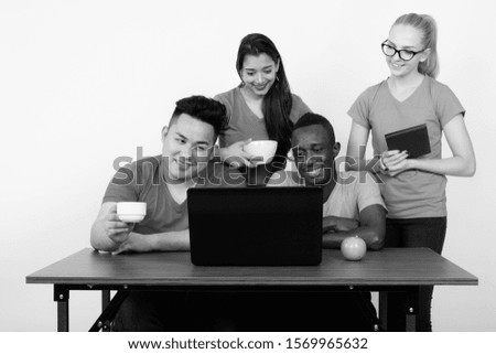 Studio shot of happy diverse group of multi ethnic friends smiling and using laptop while having coffee with green apple on wooden table together