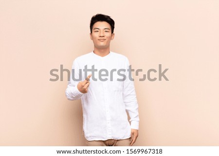 young chinese man looking proud, confident and happy, smiling and pointing to self or making number one sign against flat color wall