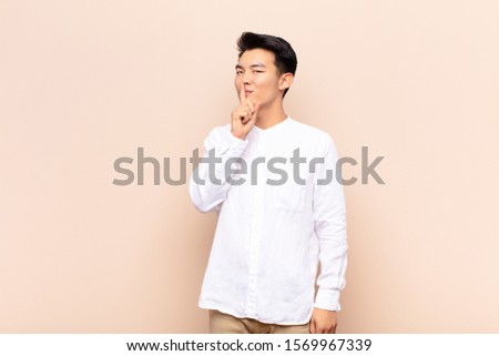 young chinese man asking for silence and quiet, gesturing with finger in front of mouth, saying shh or keeping a secret against flat color wall