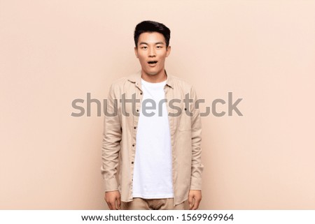 young chinese man looking very shocked or surprised, staring with open mouth saying wow against flat color wall