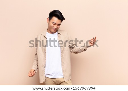 young chinese man smiling, feeling carefree, relaxed and happy, dancing and listening to music, having fun at a party against flat color wall