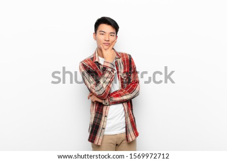 young chinese man looking serious, thoughtful and distrustful, with one arm crossed and hand on chin, weighting options against flat color wall