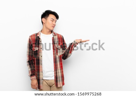 young chinese man feeling happy and smiling casually, looking to an object or concept held on the hand on the side against flat color wall