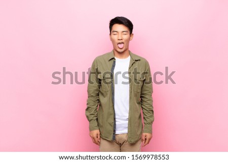 young chinese man with cheerful, carefree, rebellious attitude, joking and sticking tongue out, having fun against flat color wall