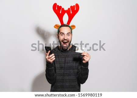 Image of happy young man in Christmas reindeer antlers headband holding smartphone and credit card isolated over gray background