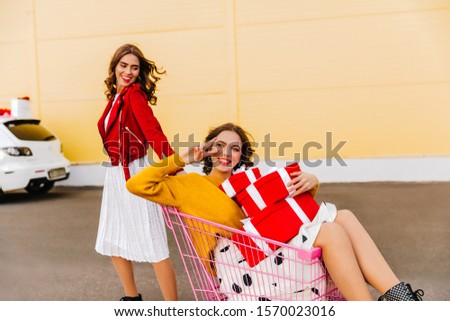 Two adorable girls posing on the street and smiling. Outdoor photo of graceful young woman in white dress chilling with friend.