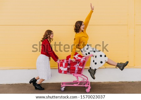Positive young woman in vintage skirt sitting in shopping cart. Two blithesome girls fooling around during photoshoot.