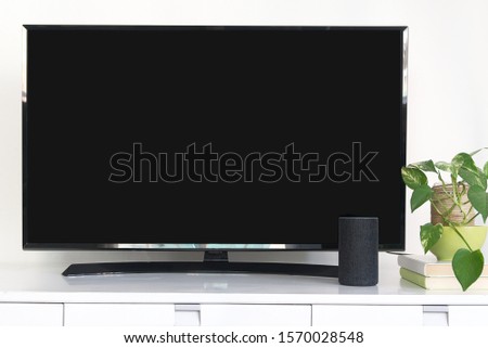 Intelligent speaker device as home automation system to control a smart television. Living room with a black screen of a tv next to some green plants.