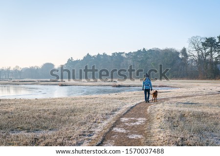 Unidentified woman with hat walks the dog on a curved path. It is early in the morning on a beautiful winter's day. The photo was taken in the Markdal nature reserve, south of the Dutch city of Breda.