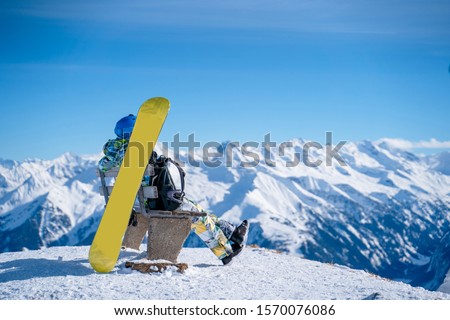 Photo from back of athlete man sitting on bench next to snowboard in ski resort.