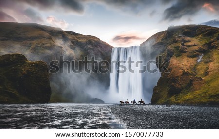 Impressively beautiful nature of Iceland during sunset. Skogafoss waterfall is one famous natural landmark and travel destination place of Iceland.  Tourists ride horses near famouse waterfall. Royalty-Free Stock Photo #1570077733