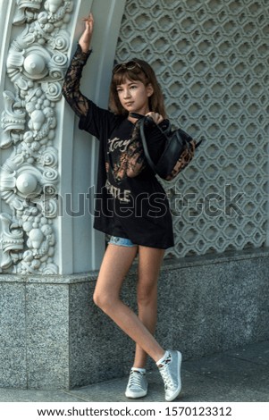 A beautiful girl in fashionable clothes stands against the wall. Portrait of a girl in black, with a backpack