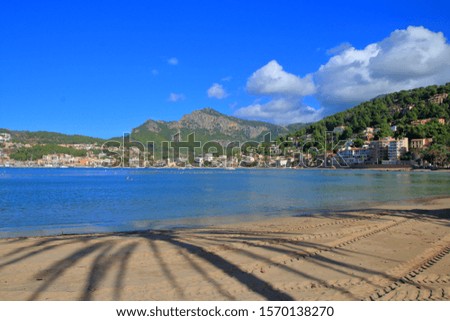 Photo taken on the island of Palma de Mallorca. The picture shows the shadows of palm trees on the sand of a beach in a picturesque bay. On the opposite side of the bay are visible a small town,