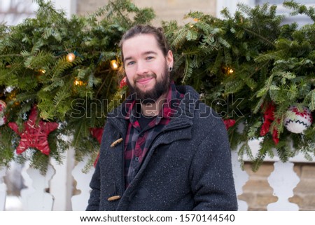 Frontal view of smiling young bearded man in warm clothes and hair tied back standing in front of gallery decorated for Christmas during a grey afternoon, Quebec City, Quebec, Canada