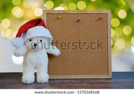 Pretty bear doll wear Christmas hat with space square board wood on bokeh background.