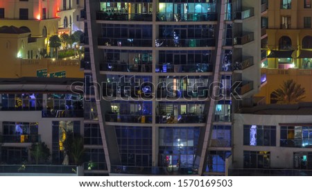 Rows of glowing windows with people in the interior of apartment building with balconies at night. Modern skyscraper with glass surface. Concept for business and modern life. Tilt up