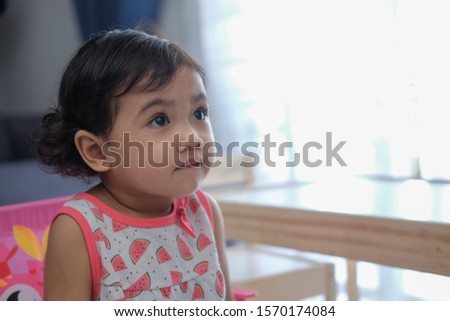 A cute little Asia baby girl with some face expression while sitting on a pink color plastic chair at home. ambient light from window.