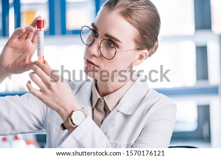selective focus of beautiful immunologist in glasses looking at test tube with virus letters 