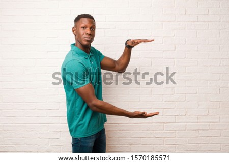 young african american black man holding an object with both hands on side copy space, showing, offering or advertising an object against brick wall