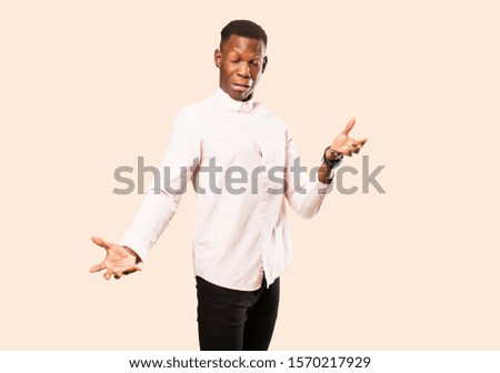 young african american black man shrugging with a dumb, crazy, confused, puzzled expression, feeling annoyed and clueless against beige wall