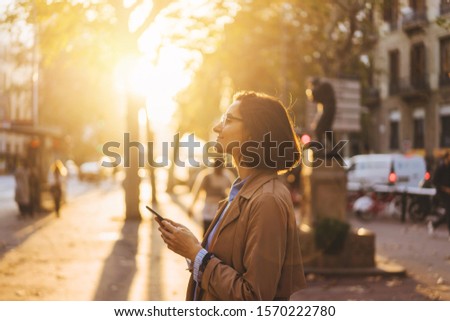 Cheerful hipster girl reading pleasant text message from best friend connected to public internet outdoors, happy woman with beautiful smile chatting on cell smartphone while relaxing outdoors