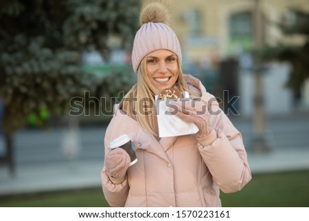 Cute young Caucasian teenage woman in beige  hat with pompon and pink mittens holding steaming cup of hot tea or coffee, outdoor in sunny winter day.