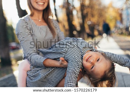 Cut view with blurred background of young woman playing with her daughter outside. Hold her in hands and smile. Child look back and lying on mother's hands. Emotional cheerful picture. Stand in park