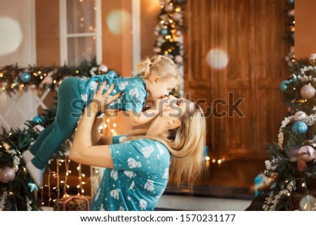 Happy blonde mother throws up her daughter baby girl in the air. Woman kiss infant. Christmas decorations and lights around.