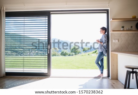 Side view of young woman with coffee standing by patio door at home. Royalty-Free Stock Photo #1570238362